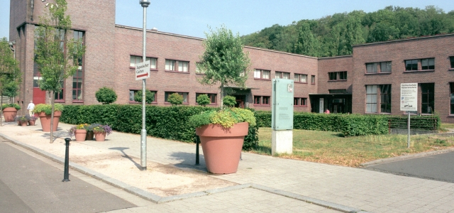 Ein Backsteinbau der Hochschule mit geschnittenen Hecken und mehreren bepflanzten kleinen und großen Blumenvasen.
