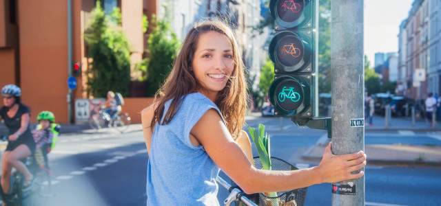 junge Frau mit Fahrrad und Einkaufskorb wartend vor einer Fahrradampel lacht in die Kamera