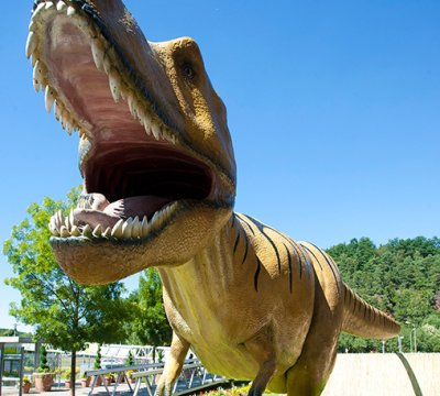 Man steht wenige Meter frontal vor der Statue des Tyranosaurus rex. 
