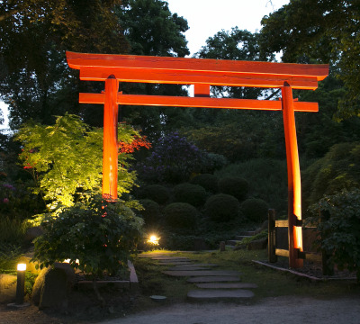 Aufnahme des illuminierten Japanischen Gartens in der Dämmerung. In der Mitte sieht man einen roten Durchgang aus Holz.
