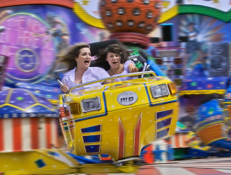 Zwei Besucherinnen werden auf einem modernen Karusell durchgeschüttelt. Mit wehenden Haaren und geöffneten Mündern, sieht man ihnen den Spaß am Nervenkitzel an.