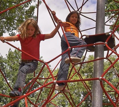 Zwei Mädchen klettern in dem mit roten Seilen bespannten Klettergerüst. 