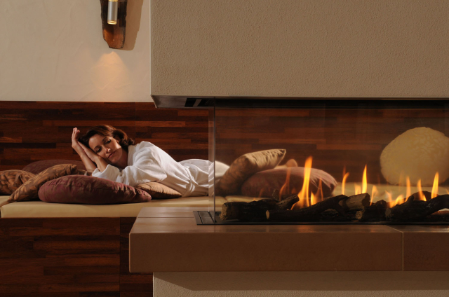 Eine Frau liegt im Bademantel auf eien Relaxzone mit Kissen und schaut einem Kaminfeuer zu. © monte mare Unternehmensgruppe