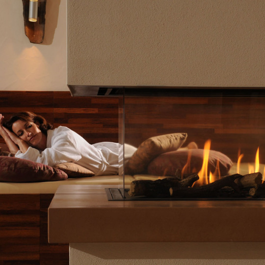 Eine Frau liegt im Bademantel auf eien Relaxzone mit Kissen und schaut einem Kaminfeuer zu.