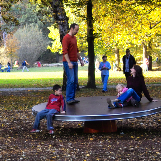 Eine Familie sitzt und steht gemeinsam auf einem drehenden Kreisel im Volkspark.