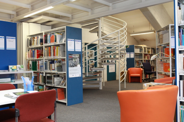 Eine einladende, kleine Bibliotheksecke mit vielen Sesseln als bequeme Sitzgelegenheiten. Über eine Wendeltrappe kann das nächste Stockwerk erreicht werden. © Hochschule Kaiserslautern