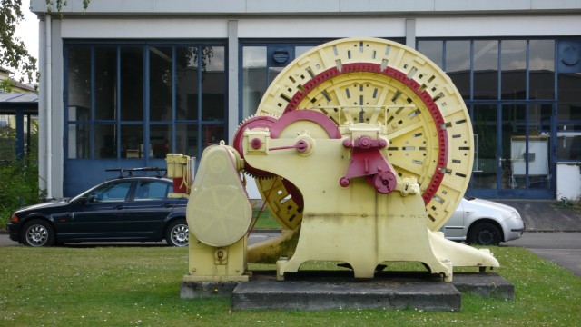 Ein großer, mit Zahnrädern und Seilen verknüpfter, alter Motor steht vor Laborgebäuden auf einer Wiese. © Hochschule Kaiserslautern