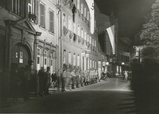 Am Rand der Straße stehen Soldaten in einer Reihe. Über ihnen weht die Flagge mit dem Symbol des Nationalsozialismus - dem Hakenkreuz. © Stadtarchiv
