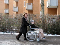 Eine ältere Dame im Rollstuhl mit einer Decke über ihren Knieen wird von einer Angehörigen über einen Weg gefahren.