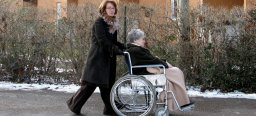 Eine ältere Dame im Rollstuhl mit einer Decke über ihren Knieen wird von einer Angehörigen über einen Weg gefahren.