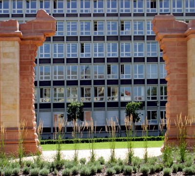 Im Vordergrund sieht man den Synagogenplatz mit Denkmal - zwei Säulen links und rechts. Im Hintergrund ein moderneres Gebäude mit großer Fensterfront.