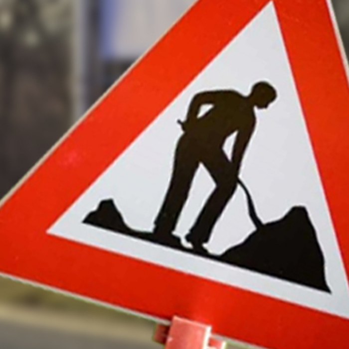 Ein Baustellenschild ragt plakativ in das Foto hinein, während sich im Hintergrund eine Straße befindet. 