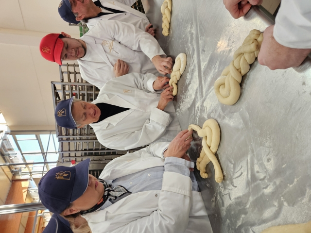 Angeleitet von Produktionsleiter Kai Drumm durfte sich die Gruppe um OB Kimmel (links Veronika Pommer, Regionalleiterin IHK) auch am Flechten eines Hefezopfes versuchen. © Stadt Kaiserslautern