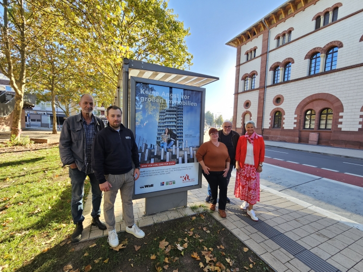 Fotokünstler Thomas Brenner und Wall-Vertriebsleiter Kevin Lorson zusammen mit Constanze Augustin und Heiko Elmenthaler von der städtischen Schwerbehindertenvertretung und Oberbürgermeisterin Beate Kimmel. Die Gruppe steht neben einem der Plakate an einer der Bushaltestellen in der Burgstraße. Rechts im Bild die Fruchthalle. © Stadt Kaiserslautern