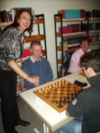 Drei Schachspieler, die an zwei Schachbrettern aus Holz sitzen. Der weiße Spieler hat seinen Eröffnungszug gemacht und den Bauer von E2 auf E4 gesetzt. Die Frau Bürgermeisterin steht am Tisch und würde am liebsten selber mitspielen.