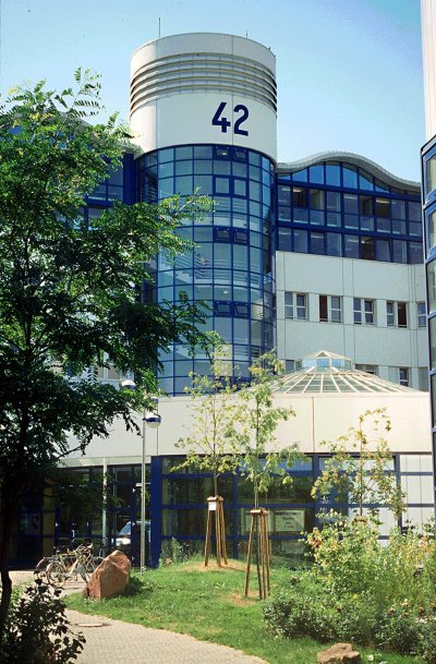 Der Turm in der Mitte des Bildes trägt die Gebäudenummer 42 und besteht zum größten Teil aus Fenstern mit blauen Rändern. Der Eingang davor liegt im Grünen und einige Fahrräder sind dort abgestellt. 