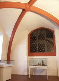 Ein Ausstellungsraum des Wadgasserhofs, der mit weißen Wänden und terrakotta farbenen Bögen an der gebogenen Decke ein kappelartiges Aussehen besitzt. Einige Exponate in Glasvitrinen sind aufgestellt.