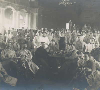 Krankenschwestern in weißen Kitteln und Haube mit dem roten Kreuz darauf, sowie Ärzte und Helfer in Kitteln sitzen im umgebauten Festsaal. 