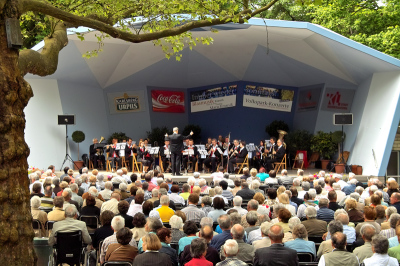 Im blauen Pavillon im Volkspark spielt ein Blasinstrumentorchester während eine große Anzahl an Zuhörern gekommen sind um zu lauschen.