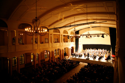 Nur die Bühne des großen Saals mit dem Orchester wird beleuchtet, der Rest ist in Dämmerlicht getaucht. Das Publikum lauscht andächtig und verfolgt die Bewegungen des Dirigenten. © Stadt Kaiserslautern