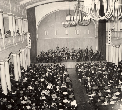 Eine schwarz-weiß Fotografie des großen Saals mit Orchesteraufführung.