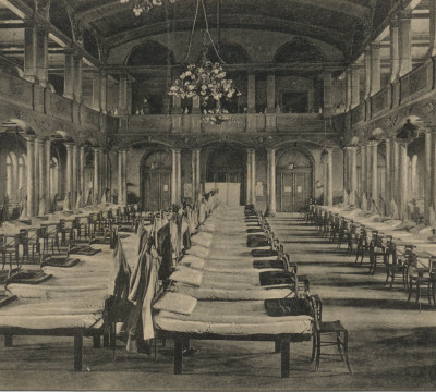 Eine schwarz-weiß Fotografie, die den großen Festsaal zeigt. Mehrere Reihen an Lazarettbetten wurden aufgestellt, davor jeweils ein Stuhl. Die festlichen Kronleuchter an der Decke ergeben mit der Lazarettumgebung ein bizarres Bild.