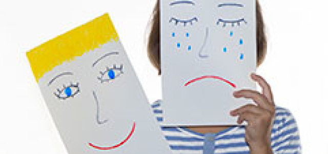 Mädchen hält ein Blatt vor das Gesicht, auf dem ein weinendes Gesicht gezeichnet ist. Die andere Hand hält ein Blatt mit einem fröhlichen Gesicht.