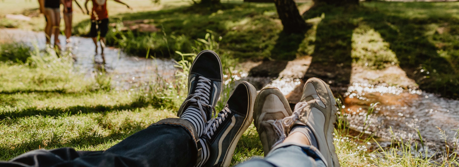 Bachlauf auf dem Gelände der Gartenschau im Sommer. Im Vordergrund liegen zwei Personen im Gras. Sehen kann man lediglich die Beine und Füße. Im Hintergrund sieht man Kinder im Wasser spielen.