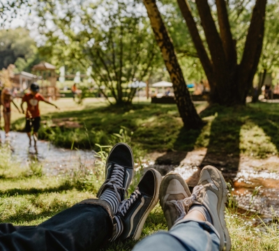 Bachlauf auf dem Gelände der Gartenschau im Sommer. Im Vordergrund liegen zwei Personen im Gras. Sehen kann man lediglich die Beine und Füße. Im Hintergrund sieht man Kinder im Wasser spielen.