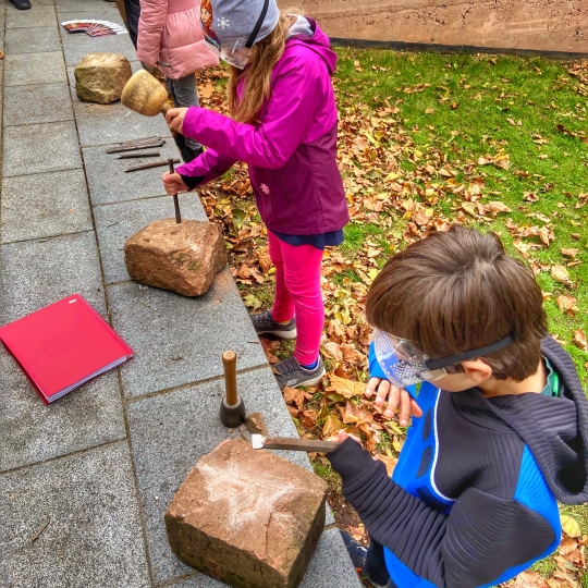 Kinder mit Schutzbrille, die an der Kaiserpfalz Steinblöcke mit Hammer und Meißel bearbeiten. 