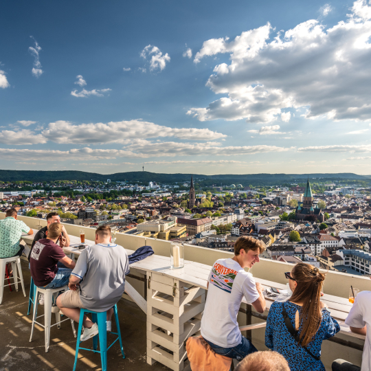 Menschen sitzen auf der Dachterrasse der Sommerlounge des Twenty One. Im Hintergrund sieht man Kaiserslautern.