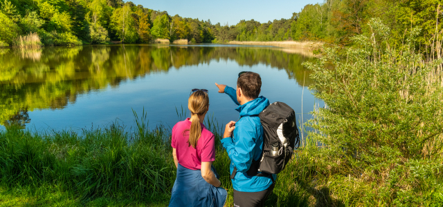 Eine Frau und ein Mann mit einem Rucksack auf dem Rücken stehen am Vogelwoog und schauen auf den See.