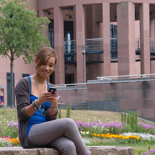 Aufnahme vor dem Pfalztheater. Eine Frau sitzt auf einem Stein und schaut auf ihr Smartphone.