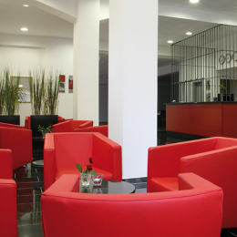 The lobby is decorated in red and black tones. The red leather chairs invite you to linger and in the background the barred reception can be seen.