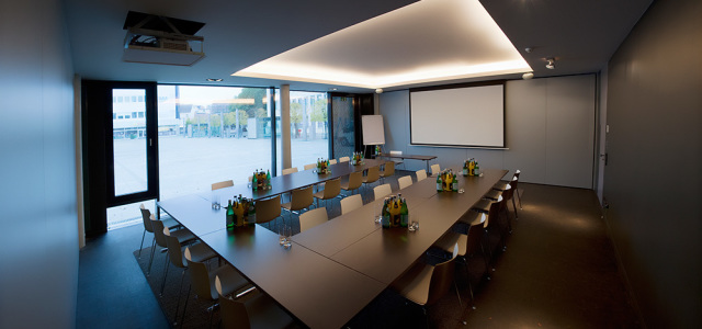 The tables of the conference room are arranged in a U-shape. There are several islands of drinks and glasses on the tables. A whiteboard and a flipchart are available for conferences. The room is on the first floor of the hotel.