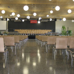 In der großen Veranstaltungshalle sind Holzstühle in Reihe zusammengestellt und zeigen Richtung Bühne.