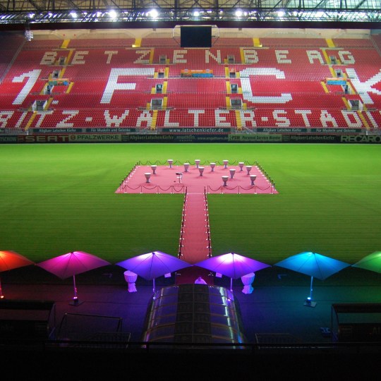 Blick auf das Spielfeld im Fritz-Walter-Stadion mit aufgebauter Veranstaltung. Im Vordergrund beleuchtete Sonnenschirme. Im Mittelkreis Stehtische.